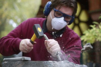 Mann im dunkelrotem Pulli, mit Mundschutz, Augen- und Gehörschutz, der mit Hammer und Meißel einen Stein bearbeitet.