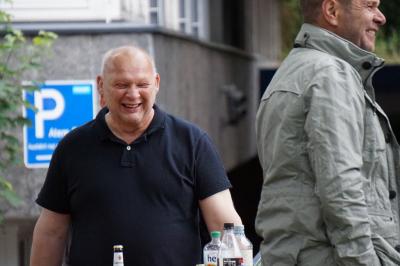 Lachender, etwa 45jähriger Mann in blauem T-Shirt mit Halbglatze, im Vordergrund ein weiterer lachender Mann von der Seite (in beiger Jacke), im Hintergrund ein Gebäude und ein "Parkplatz"-Schild Lachender, etwa 45jähriger Mann in blauem T-Shirt mit Halbglatze, im Vordergrund ein weiterer lachender Mann von der Seite (in beiger Jacke), im Hintergrund ein Gebäude und ein "Parkplatz"-Schild
