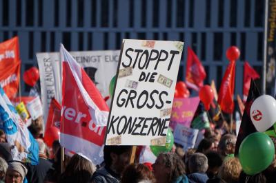 Menschen, Fahnen, dazu ein Papp-Plakat mit der Aufschrift "Stoppt die Gross-Konzerne". Menschen, Fahnen, dazu ein Papp-Plakat mit der Aufschrift "Stoppt die Gross-Konzerne".