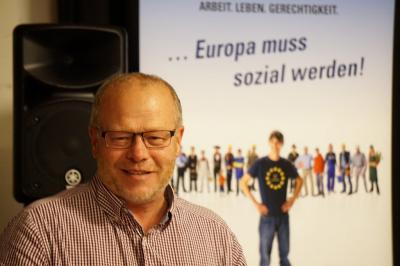 Ein freundlich lächelnder Mann mit Brille, hoher Stirn und Dreitagebart in kariertem Hemd. Im Hintergrund ein Plakat mit der Aufschrift &quot;Europa muss sozial werden!&quot;