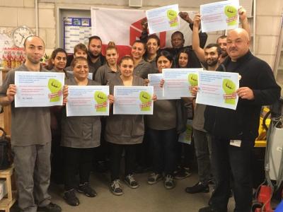 16 Menschen, Männer und Frauen, in grauer Arbeitskleidung stehen im Halbkreis vor einer Fahne der IG BAU und halten Plakate mit Tarifforderungen in die Kamera.