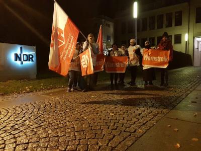 Etwa ein Dutzend Menschen, überwiegend mit rot-weißen Streikplakaten und Gewerkschaftsfahnen auf einer gepflasterten Auffahrt. Im Hintergrund der beleuchtete Schriftzug "NDR". Es ist noch dunkel, im Hintergrund leuchtet eine Straßenlaterne. Etwa ein Dutzend Menschen, überwiegend mit rot-weißen Streikplakaten und Gewerkschaftsfahnen auf einer gepflasterten Auffahrt. Im Hintergrund der beleuchtete Schriftzug "NDR". Es ist noch dunkel, im Hintergrund leuchtet eine Straßenlaterne.
