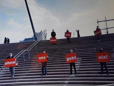 Ein halbes Dutzend Reinigungskräfte mit roten Schildern auf einer großen Treppe, im Hintergrund die Elbphilharmonie.