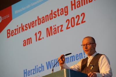 Matthias Maurer vor hell erleuchteter Leinwand mit der Aufschrift "Bezirksverbandstag Hamburg am 12. März 2022 Matthias Maurer vor hell erleuchteter Leinwand mit der Aufschrift "Bezirksverbandstag Hamburg am 12. März 2022