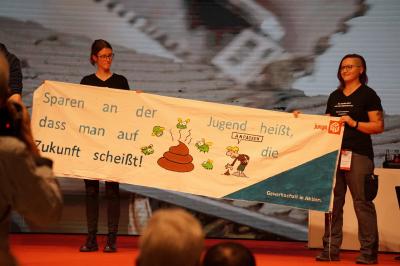 Zwei junge Gewerkschafterinnen mit einem Transparent auf der Bühne. Darauf abgebildet u.a. ein Haufen Scheiße und der Slogan: Sparen an der Jugend heißt, dass man auf die Zukunft scheißt.