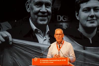 Robert Feiger vor einem übergroßen Schwarz-Weiß-Foto, auf dem er mit Anderen auf einer Demonstration zu sehen ist. Feiger trägt ein weißes Hemd und den obligatorischen Delegiertenausweis am roten Band und steht am Rednerpult. Robert Feiger vor einem übergroßen Schwarz-Weiß-Foto, auf dem er mit Anderen auf einer Demonstration zu sehen ist. Feiger trägt ein weißes Hemd und den obligatorischen Delegiertenausweis am roten Band und steht am Rednerpult.