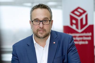 Carsten Burckhardt in blauem Jackett und weißem Hemd, im Hintergrund ein BAU-Banner Carsten Burckhardt in blauem Jackett und weißem Hemd, im Hintergrund ein BAU-Banner