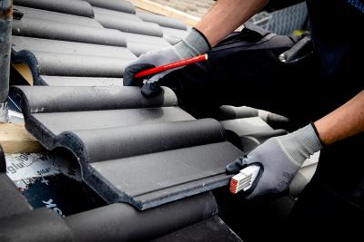 Ein schwarzer Dachziegel in den Händen eines Mannes. Er trägt Handschuhe und hält außerdem einen Zollstock und einen Bleistift. Ein schwarzer Dachziegel in den Händen eines Mannes. Er trägt Handschuhe und hält außerdem einen Zollstock und einen Bleistift.