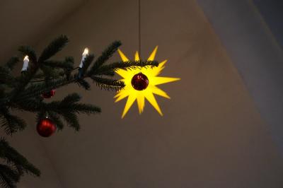 Teile eines Weihnachtsbaumes mit Kerzen und roten Christbaumkugeln. Eine Kugel hängt vor einem gelben, beleuchteten Herrnhuther Stern.