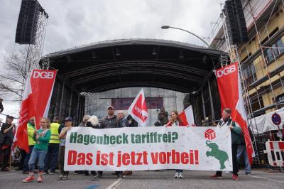 Etwa ein dutzend Menschen vor ver.di-Fahnen und DGB-Bannern tragen ein Transparent mit der Aufschrift "Hagenbeck tariffrei? Das ist jetzt vorbei!". Im Hintergrund eine große Bühne.
