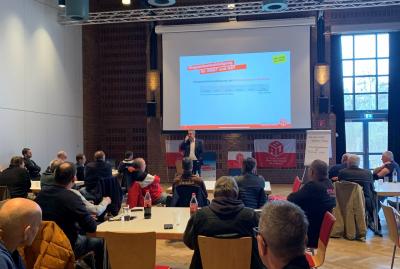 Größerer Saal mit Leinwand, auf die eine Powerpoint-Präsentation projiziert wird. Im Vordergrund einige Menschen auf Stühlen, die auf den Referenten Carsten Burckhardt blicken. Größerer Saal mit Leinwand, auf die eine Powerpoint-Präsentation projiziert wird. Im Vordergrund einige Menschen auf Stühlen, die auf den Referenten Carsten Burckhardt blicken.