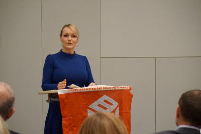 Eine blonde Frau im blauen Kleid an einem Rednerpult. Vorne am Pult hängt eine rote Fahne mit der Aufschrift "Industriegewerkschaft Bauen-Agrar-Umwelt". Eine blonde Frau im blauen Kleid an einem Rednerpult. Vorne am Pult hängt eine rote Fahne mit der Aufschrift "Industriegewerkschaft Bauen-Agrar-Umwelt".