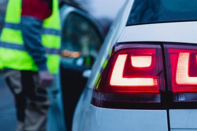 Das linke Rücklicht eines Autos in Nahaufnahme, im Hintergrund schemenhaft eine Person in Warnweste, die die Fahrertür öffnet. Das linke Rücklicht eines Autos in Nahaufnahme, im Hintergrund schemenhaft eine Person in Warnweste, die die Fahrertür öffnet.