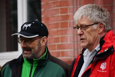 Zwei Männer, die in dieselbe Richtung blicken. Einer der Männer trägt eine schwarze Kappe und ein gründes Oberteil, der andere haut graue Haare und trägt eine rote IG BAU-Jacke.