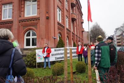 Ein Mann in roter IG BAU-Jacke spricht vor einem Transparent im Garten eines roten Backsteinhauses. Knapp ein dutzend Menschen hören zu.