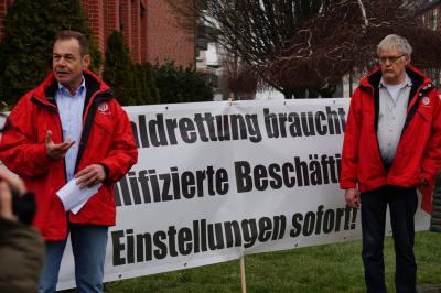 Zwei Männer in roten IG BAU-Jacken vor einem Transparent. Schwarze Schrift auf weißem Stoff: "Waldrettung braucht qualifizierte Beschäftigte - Einstellungen sofort!"