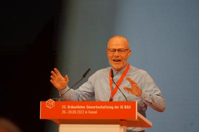 Ein etwa 60 Jahre alter Mann mit Brille und blau-weiß kariertem Hemd steht an einem Rednerpult und spricht. Beide Hände sind erhoben, am Rednerpult steht in weiß auf rotem Untergrund "23. Ordentlicher Gewerkschaftstag der IG BAU, 26.-29.09.2022 in Kassel". Ein etwa 60 Jahre alter Mann mit Brille und blau-weiß kariertem Hemd steht an einem Rednerpult und spricht. Beide Hände sind erhoben, am Rednerpult steht in weiß auf rotem Untergrund "23. Ordentlicher Gewerkschaftstag der IG BAU, 26.-29.09.2022 in Kassel".